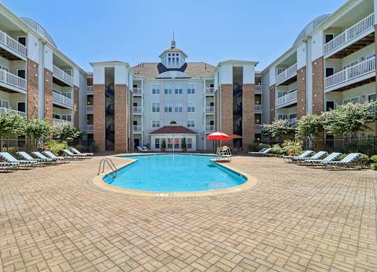 A large swimming pool surrounded by a brick patio and lounge chairs in front of apartment buildings.