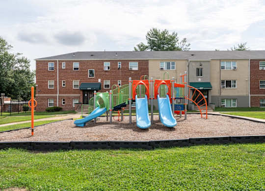 A playground with a green slide and orange safety barriers in front of a brick building.