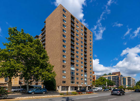 A tall brown building with many windows is in the middle of a street with cars and trees.