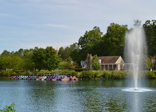 A fountain in the middle of a lake with a house and trees in the background.