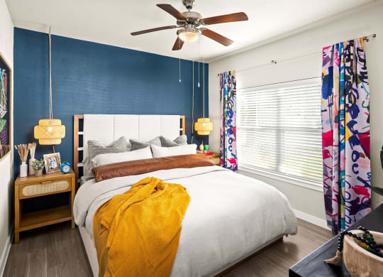 bedroom with blue accent wall behind bed. ceiling fan with handing accent lighting. bright curtains and moody decor.