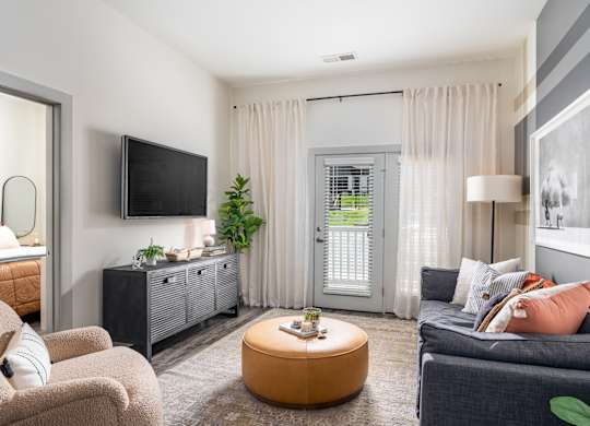 A living room with a grey couch, a brown ottoman, and a flat screen TV mounted on the wall.