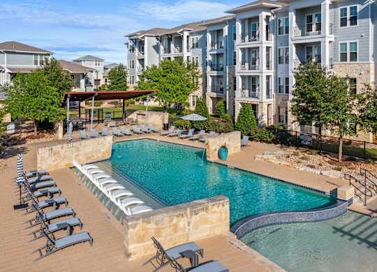 A swimming pool surrounded by sun loungers in front of apartment buildings.