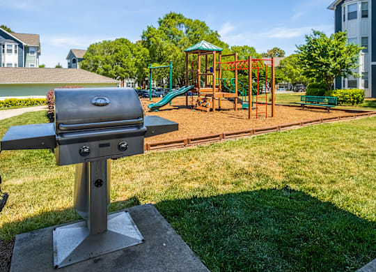 A barbecue grill is in the foreground of a park with a playground in the background.
