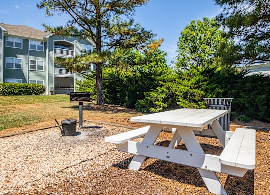 A white picnic table is in the foreground of a gravel area with a tree and apartment building in the background.