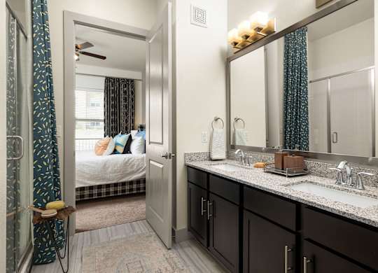 A bathroom with a mirror, sink, and a bedroom with a bed and a window.