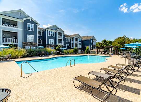 A pool surrounded by lounge chairs and umbrellas in front of apartment buildings.