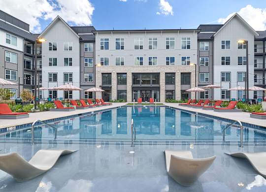 A large pool with lounge chairs and umbrellas in front of a multi-story apartment building.