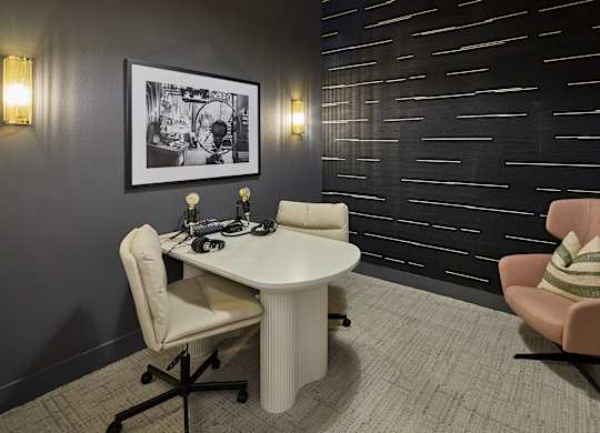A black and white picture hangs on a dark wall above a white table with a laptop and a chair with a striped pillow on it.