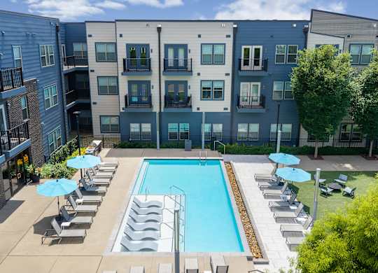 A swimming pool surrounded by lounge chairs and umbrellas in front of a multi-story apartment building.
