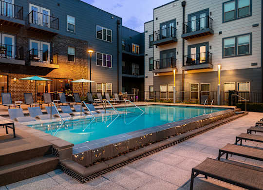 A swimming pool surrounded by lounge chairs and umbrellas in front of apartment buildings.