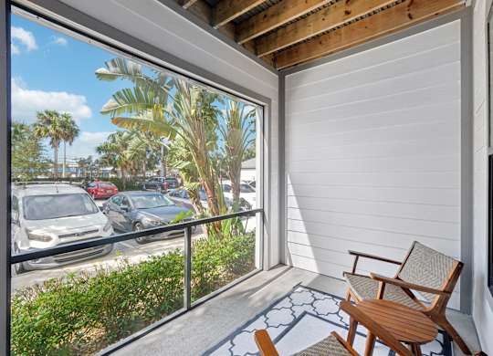 A wooden chair is on a patio with a view of a parking lot and palm trees.