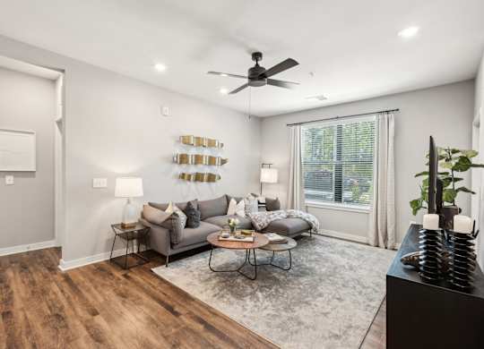 A living room with a grey couch, a coffee table, a ceiling fan, and a large window.