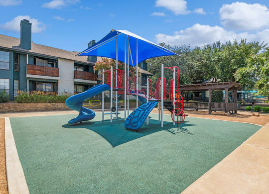 Community Playground with a Slide and Blue Canopy at Bridges at Deer Run Apartments in Dallas, TX.
