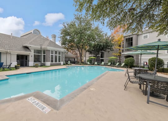 2-Bedroom Apartments in Lewisville, TX - Bridges at Oakbend - Outdoor Pool With Lounge Chairs, Patio Tables With Umbrellas, And Clubhouse Surrounded By Trees.