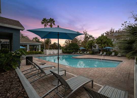 Westside Jacksonville, FL, Apartments Near Oakleaf Town Center - Westland Park - A Swimming Pool Area Features Lounge Chairs, Blue Umbrellas, And Palm Trees.