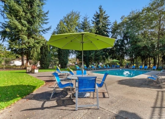 Federal Way, WA, Apartments Near Costco - Sunset Three Twenty - A Swimming Pool With Blue Lounge Chairs, Concrete Deck, Green Umbrella Seating Area, And Tall Evergreen Trees.