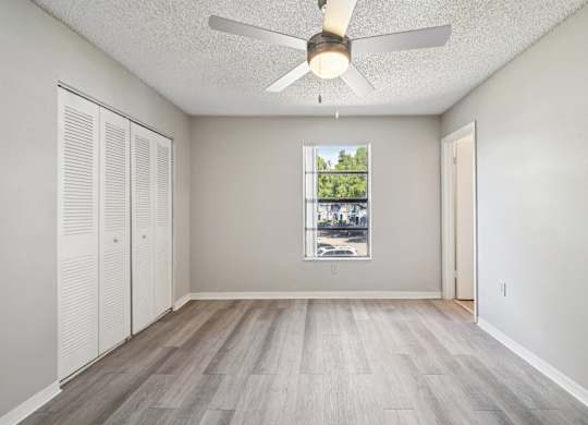 A room with a ceiling fan and a window showing a view of trees and buildings outside.