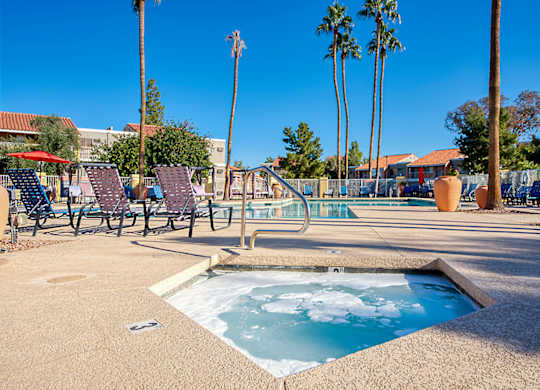 A hot tub sits in the middle of a patio with chairs and palm trees in the background.