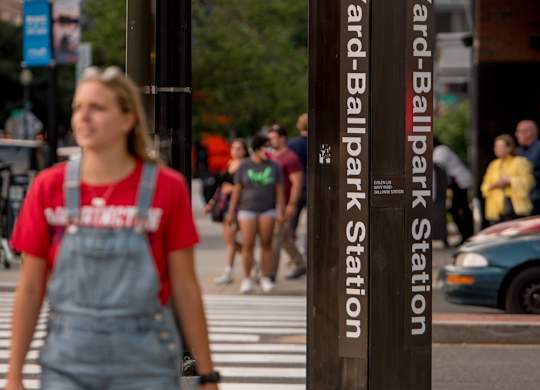 A woman in a red shirt and denim overalls is walking across the street at Navy Yard Ballpark Station.