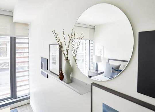 A round mirror hangs on a wall above a shelf with vases and pictures.