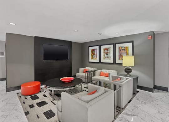 A modern living room with a grey couch, a black and white rug, a red ottoman, a black coffee table, a flat screen TV, and framed pictures on the wall.