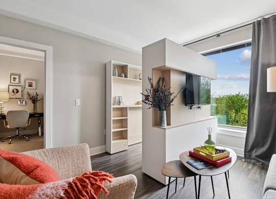 A modern living room with a white couch, a coffee table, and a large window with a view of the trees outside.