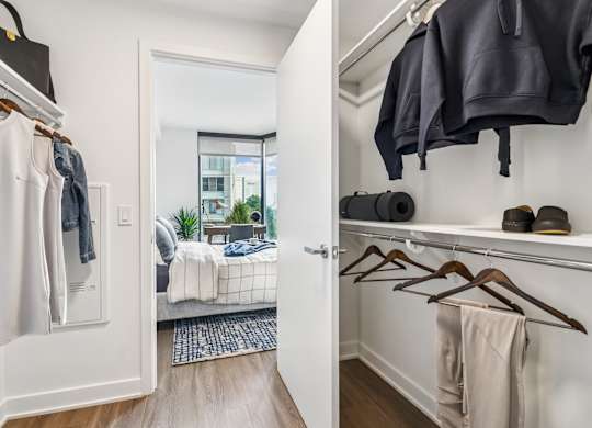 A white closet with clothes hanging and a bedroom in the background.