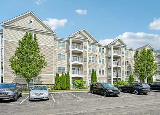 A row of cars are parked in a lot in front of apartment buildings.