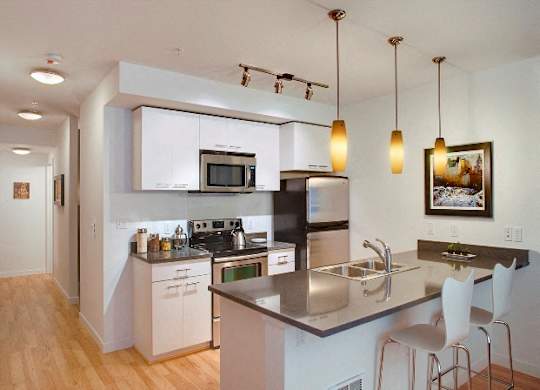 One-Bedroom Apartments In Green Lake, Seattle, WA - Circa Green Lake - Kitchen With Hardwood Floor, Stainless Steel Appliances, Kitchen Bar, Pendant Lights, Quartz Countertops, And White Storage Cabinets.