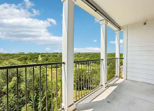 A balcony with white columns and a black railing overlooks a green landscape.