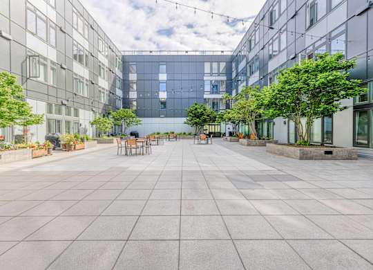 Green Lake, Seattle, WA, Apartments For Rent - Circa Green Lake - Courtyard With Picnic Tables And Chairs, Cafe Lights, Plants, Trees, And Surrounding Apartment Buildings.