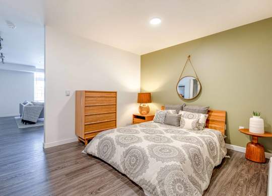 Studio Apartments In Green Lake, Seattle, WA - Circa Green Lake - Spacious Bedroom With Hardwood Floor, Bed, Round Mirror, Nightstand with Lamp, Round Side Table, Dresser, and View of Living Room.