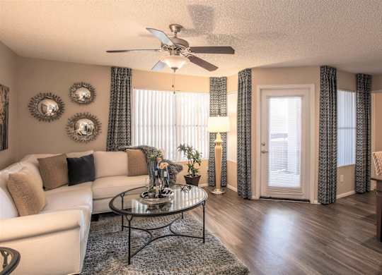 Dog-Friendly Apartments in Ahwatukee, AZ - Pacific Bay Club - Living Room with Wood-Style Plank Flooring, Area Rug, White Sectional Couch, Circular Glass Coffee Table, Dining Area with Table and Chairs, Decorative Art, Standing Lamp, Ceiling Fan, Windows with Curtains, Door with Access to Patio or Balcony.