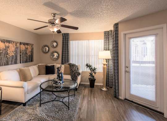 Apartments in Ahwatukee, Foothills, AZ - Pacific Bay Club - Living Room with Wood-Style Plank Flooring, Stylish Decor, Ceiling Fan, Large Window, and Door to the Private Patio