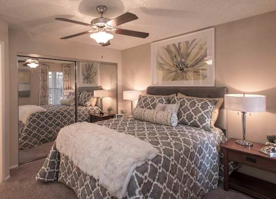 One and Two-Bedroom Apartments in Ahwatukee, AZ - Pacific Bay Club - Bedroom with Mirrored Closet Doors, Carpet Floors, Ceiling Fan, Cozy Bed, Stylish Decor, and Door to Bathroom with Wood-Style Plank