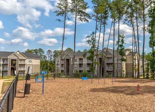 A playground with a sandy area and a few trees in front of apartment buildings.