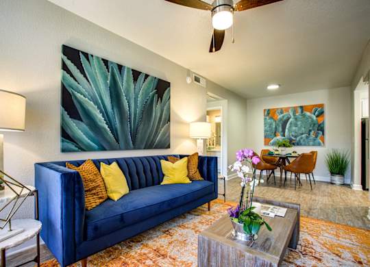 South Phoenix Apartments for Rent - Village at Lakewood - Living Room with Wood-Style Flooring, Orange Area Rug, Blue Sofa, Wooden Coffee Table, Ceiling Fan, and Wall Art