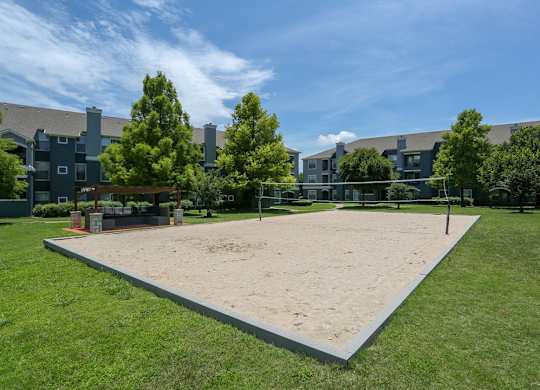 A sand volleyball court is surrounded by grass and trees.
