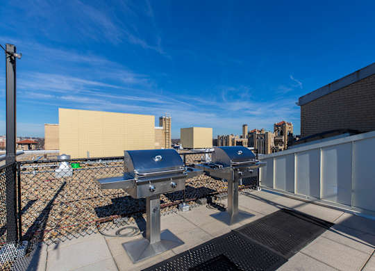 A grill is on a patio with a white fence and a building in the background.