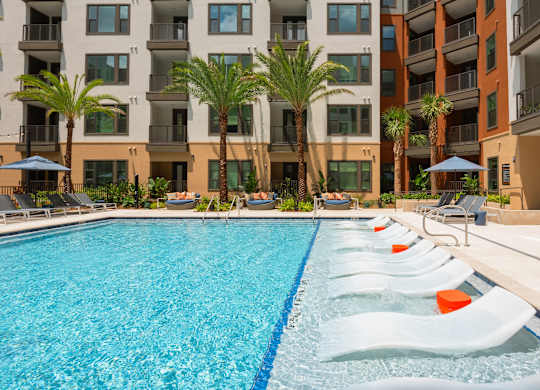 A swimming pool with lounge chairs and palm trees in front of a multi-story apartment building.