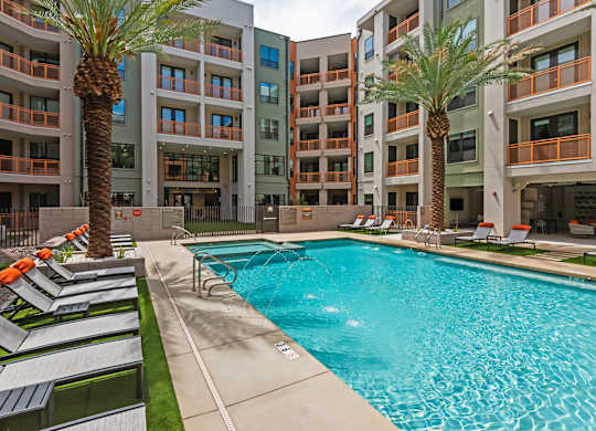 A swimming pool surrounded by palm trees and lounge chairs in front of apartment buildings.