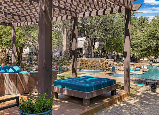 A wooden pergola over a sunbed with a pool in the background.