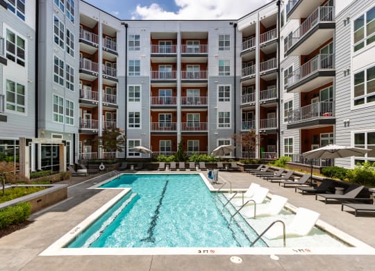 an outdoor swimming pool with lounge chairs and umbrellas in front of an apartment building  at Abberly Noda Vista Apartment Homes, North Carolina