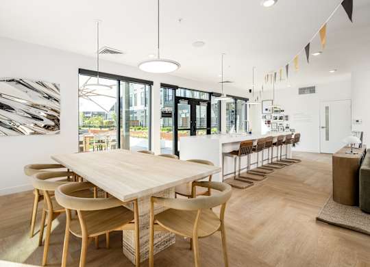 A long wooden table is surrounded by chairs in a white room with a large window.