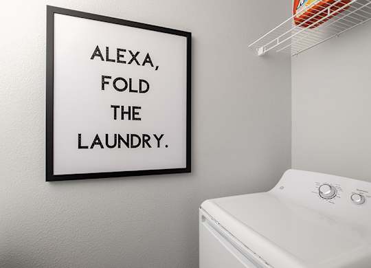 A white washing machine sits under a picture frame that says