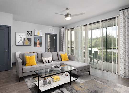 A modern living room with a grey sofa, a glass coffee table, and a ceiling fan.