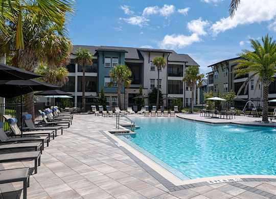 A swimming pool surrounded by lounge chairs and umbrellas in front of a row of apartment buildings.