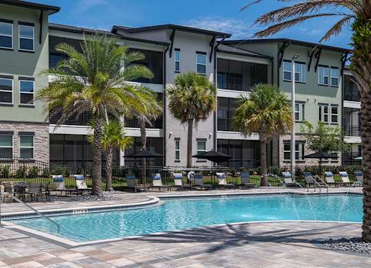 A swimming pool is surrounded by palm trees and lounge chairs in front of apartment buildings.