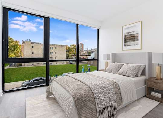 A bedroom with a large bed and a view of a building and trees outside the window.